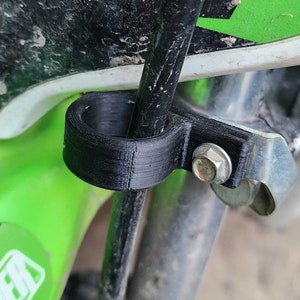 Puede incluir: Una abrazadera de plástico negro con un soporte metálico y un tornillo. La abrazadera está unida a un cable negro y se utiliza para sujetar el cable a una superficie.