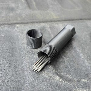 Puede incluir: Una herramienta de metal negro con un cuerpo cil&iacute;ndrico y un extremo hexagonal, que contiene varias varillas puntiagudas de color plateado. Una pieza cil&iacute;ndrica separada y a juego est&aacute; cerca. Los art&iacute;culos est&aacute;n sobre una superficie gris texturizada.