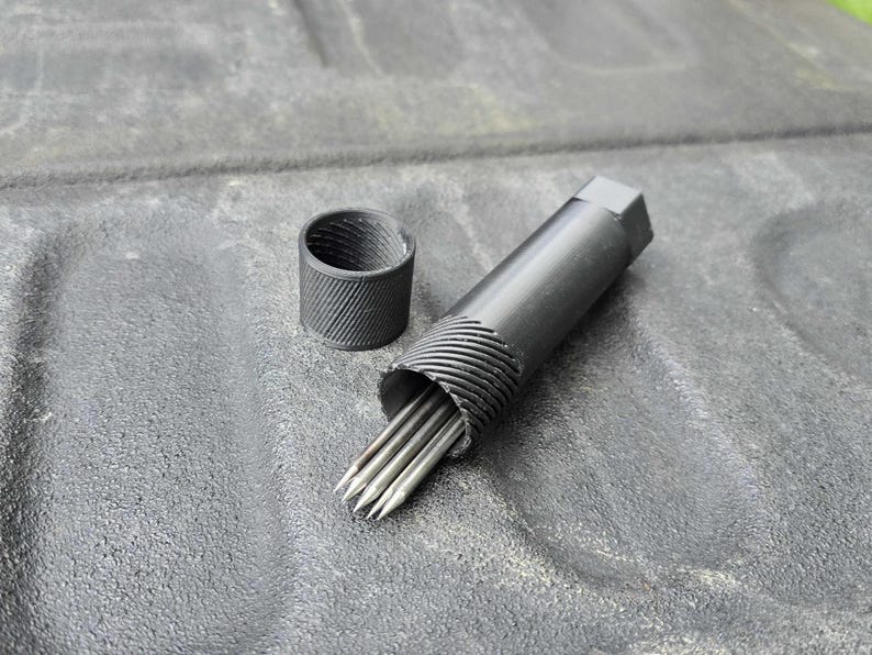 Puede incluir: Una herramienta de metal negro con un cuerpo cil&iacute;ndrico y un extremo hexagonal, que contiene varias varillas puntiagudas de color plateado. Una pieza cil&iacute;ndrica separada y a juego est&aacute; cerca. Los art&iacute;culos est&aacute;n sobre una superficie gris texturizada.