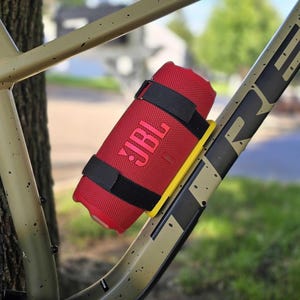 Puede incluir: Un altavoz portátil JBL cilíndrico de color rojo sujeto al cuadro de una bicicleta de montaña de color tostado con correas negras. El altavoz presenta un logotipo JBL rojo en su exterior de malla y está montado sobre un soporte de plástico amarillo.