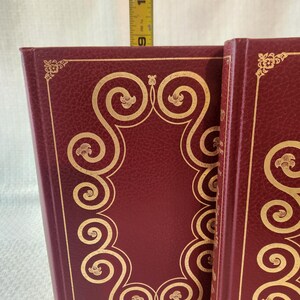 Peut inclure: Deux livres bordeaux avec des motifs de volutes en feuille d'or sur les couvertures. Un m&egrave;tre ruban jaune est plac&eacute; derri&egrave;re les livres, indiquant une hauteur d'environ 24 cm de la base jusqu'au bord sup&eacute;rieur des couvertures des livres.