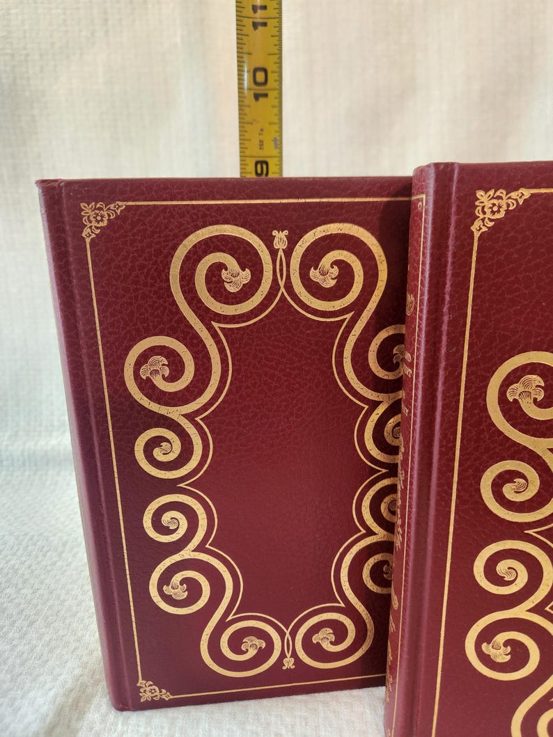 Peut inclure: Deux livres bordeaux avec des motifs de volutes en feuille d'or sur les couvertures. Un m&egrave;tre ruban jaune est plac&eacute; derri&egrave;re les livres, indiquant une hauteur d'environ 24 cm de la base jusqu'au bord sup&eacute;rieur des couvertures des livres.