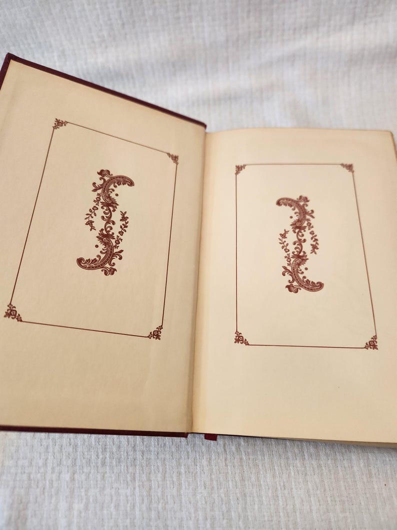 Peut inclure: Un livre &agrave; couverture rigide ouvert avec des pages de garde couleur cr&egrave;me. Chaque page pr&eacute;sente une fine bordure rectangulaire marron avec des accents d&eacute;coratifs aux coins et un motif central vertical en filigrane marron.