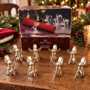 May include: A set of eight silver-plated angel napkin rings, each featuring a small angel figure holding a trumpet. The items are displayed in front of a rectangular cardboard box labeled Royal Silver 4 Angel Napkin Silver Plate.