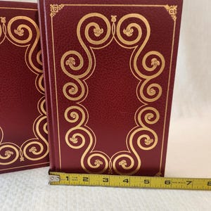 Peut inclure: Deux livres reli&eacute;s bordeaux avec des motifs de volutes en feuille d'or sur les couvertures avant. Un ruban &agrave; mesurer jaune est plac&eacute; le long du bord inf&eacute;rieur, montrant que la largeur du livre est d'environ 19 centim&egrave;tres du dos au bord.