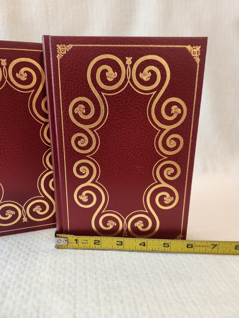 Peut inclure: Deux livres reli&eacute;s bordeaux avec des motifs de volutes en feuille d'or sur les couvertures avant. Un ruban &agrave; mesurer jaune est plac&eacute; le long du bord inf&eacute;rieur, montrant que la largeur du livre est d'environ 19 centim&egrave;tres du dos au bord.
