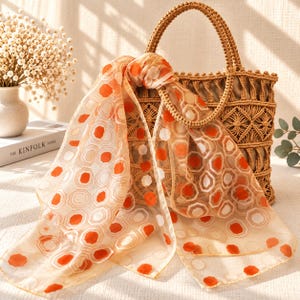 May include: A tan woven macrame handbag with two rounded handles, draped with a sheer, lightweight scarf featuring a repeating geometric pattern of orange and white octagons and thin lines on a translucent, light beige background.