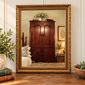 May include: A rectangular wall mirror featuring an ornate, antique gold-toned frame with intricate scrollwork and beaded detailing. The reflective glass surface displays a clear view of a room interior, including a dark wood armoire and bedding.