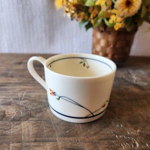 May include: White ceramic teacup with a handle, adorned with a delicate floral design in orange, green, and blue. The cup features a dark blue band near the rim and base. The cup is on a wooden surface.