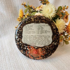 May include: A round, resin-coated coaster featuring a vintage recipe for Seafood Gumbo. The coaster has a dark brown border with embedded decorative elements and a central image of a crab. The recipe text is visible.