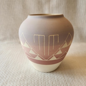 May include: A ceramic vase with a rounded body and a narrow opening. The vase features a color gradient from light purple to pink, with geometric designs in white. The base is white.