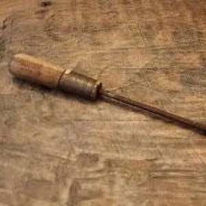 May include: A vintage hand tool featuring a cylindrical wooden handle with a weathered texture, connected to a dark metallic ferrule and a long, slender, pointed steel shaft that extends outward, showing signs of oxidation and age-related wear.