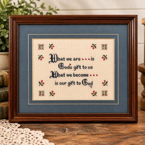 May include: A framed cross-stitch sampler featuring the text "What we are... is God's gift to us. What we become... is our gift to God" in black lettering, surrounded by small red and green floral motifs on a cream fabric background.