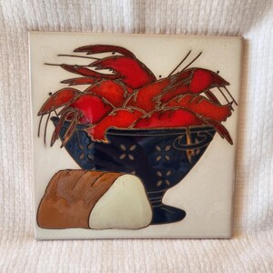 May include: Square ceramic tile featuring a vibrant illustration of red crawfish overflowing from a dark blue bowl. A loaf of bread is positioned next to the bowl. The tile has a cream-colored background and is likely for kitchen decor.