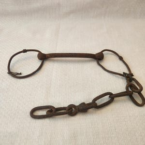 May include: A rusty brown metal horse bit with a central bar, two curved side pieces, and a chain. The bit is designed for use in equestrian activities. The metal has a weathered appearance.