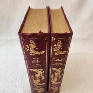 Peut inclure: Deux livres reli&eacute;s avec des couvertures textur&eacute;es rouge fonc&eacute; et des tranches dor&eacute;es. Les dos pr&eacute;sentent des motifs en volutes et des lettrages dor&eacute;s en relief. Un livre est intitul&eacute; War and Peace de Leo Tolstoy, l'autre Crime and Punishment de Fyodor Dostoevsky.