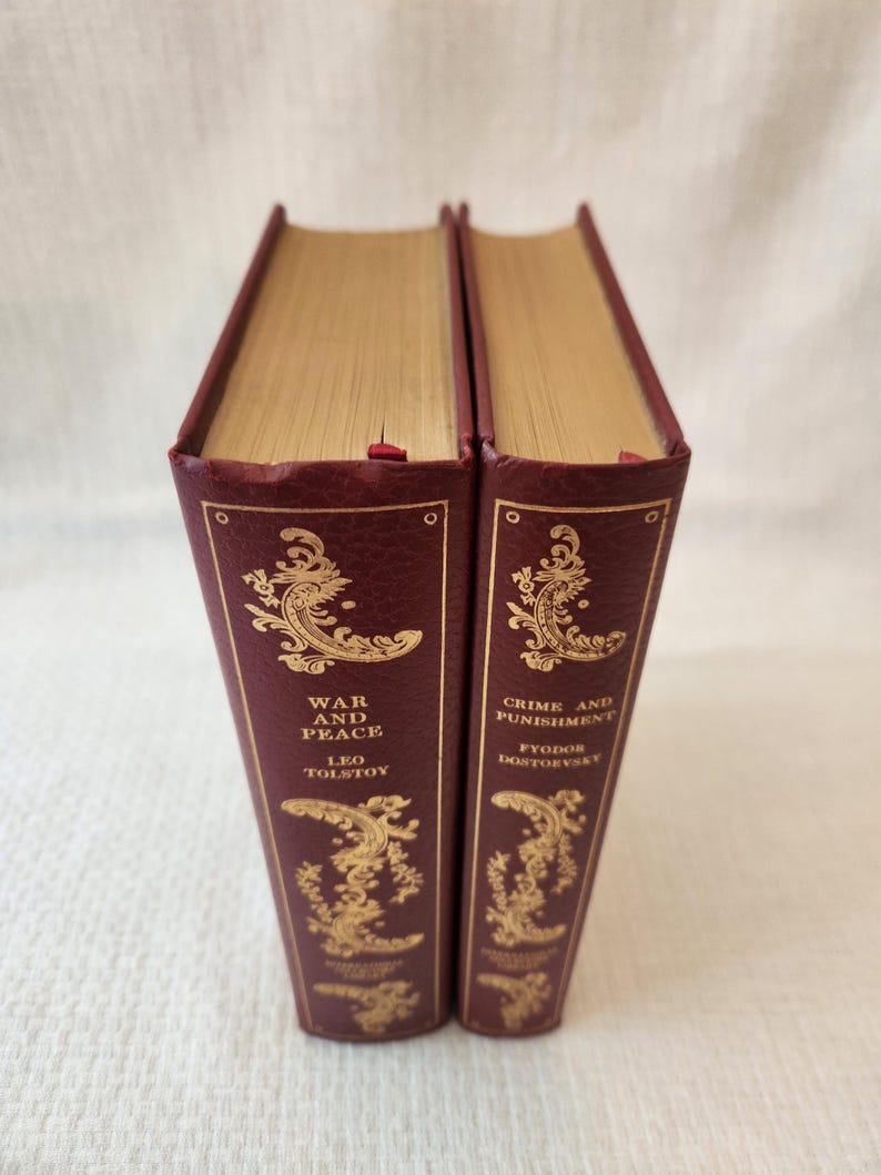 Peut inclure: Deux livres reli&eacute;s avec des couvertures textur&eacute;es rouge fonc&eacute; et des tranches dor&eacute;es. Les dos pr&eacute;sentent des motifs en volutes et des lettrages dor&eacute;s en relief. Un livre est intitul&eacute; War and Peace de Leo Tolstoy, l'autre Crime and Punishment de Fyodor Dostoevsky.