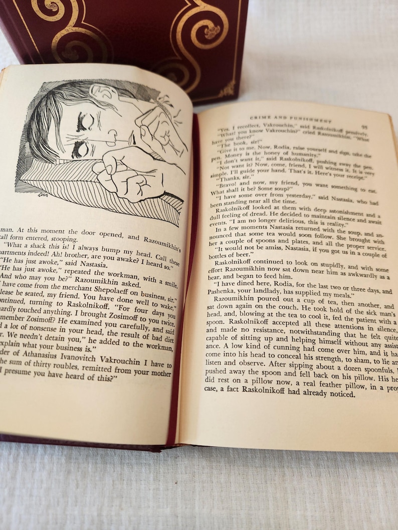 Peut inclure: Un livre reli&eacute; ouvert intitul&eacute; Crime and Punishment avec une couverture rouge fonc&eacute; et des accents d&eacute;coratifs dor&eacute;s. La page de gauche pr&eacute;sente une illustration &agrave; l'encre noire et blanche d'une personne allong&eacute;e, et la page de droite du texte.