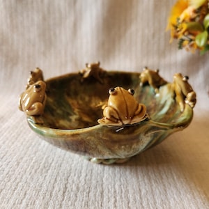 May include: A ceramic bowl with a brown and green glaze, adorned with several small, tan frog figurines around the rim. The bowl has a unique, organic shape and sits on a white surface. The frogs are detailed with visible eyes and textured skin.
