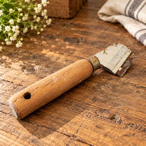 May include: A handheld paint scraper featuring a cylindrical wooden handle with a small circular hole at the end and a weathered metal blade attached by a rusted metal ferrule. The tool shows signs of wear with visible oxidation on the metal.