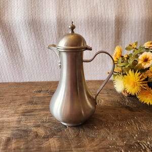 May include: A vintage silver-colored pitcher with a lid and handle, set on a wooden surface. The pitcher has a bulbous body and a slender neck, with a small spout. A bouquet of yellow flowers is in the background.