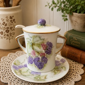 May include: A white porcelain lidded sugar bowl with two handles and a matching saucer, featuring hand-painted purple blackberry clusters and green leaves. The lid has a small blackberry-shaped knob, and the rim is accented with gold trim.
