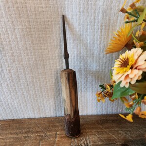 May include: A rustic, hand-crafted wooden awl with a dark, pointed metal tip. The wooden handle features a natural, two-tone finish, showcasing the wood grain. The awl is displayed on a wooden surface, with a floral arrangement in the background.