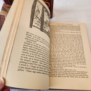 Peut inclure: Un livre reli&eacute; ouvert avec des pages couleur cr&egrave;me pr&eacute;sentant du texte imprim&eacute; en noir et une illustration au trait en noir et blanc d'une personne regardant par une fen&ecirc;tre. Le livre a une couverture bordeaux fonc&eacute; avec des motifs dor&eacute;s.