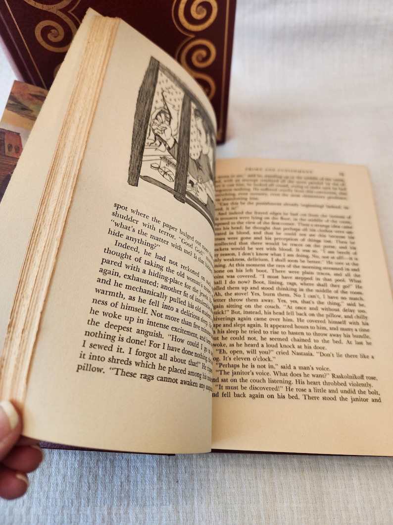 Peut inclure: Un livre reli&eacute; ouvert avec des pages couleur cr&egrave;me pr&eacute;sentant du texte imprim&eacute; en noir et une illustration au trait en noir et blanc d'une personne regardant par une fen&ecirc;tre. Le livre a une couverture bordeaux fonc&eacute; avec des motifs dor&eacute;s.