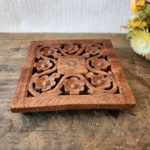 May include: A square, wooden decorative piece with intricate floral carvings. The brown wood has a natural finish, and the design features symmetrical flower and vine patterns. The piece is likely a coaster or small decorative item.