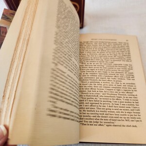 Peut inclure: Un livre reli&eacute; ouvert avec des pages couleur cr&egrave;me et du texte imprim&eacute; en noir. Le c&ocirc;t&eacute; gauche montre des pages floues en &eacute;ventail, tandis que le c&ocirc;t&eacute; droit affiche une page de texte claire tir&eacute;e de Crime et Ch&acirc;timent. Une couverture rouge fonc&eacute;.