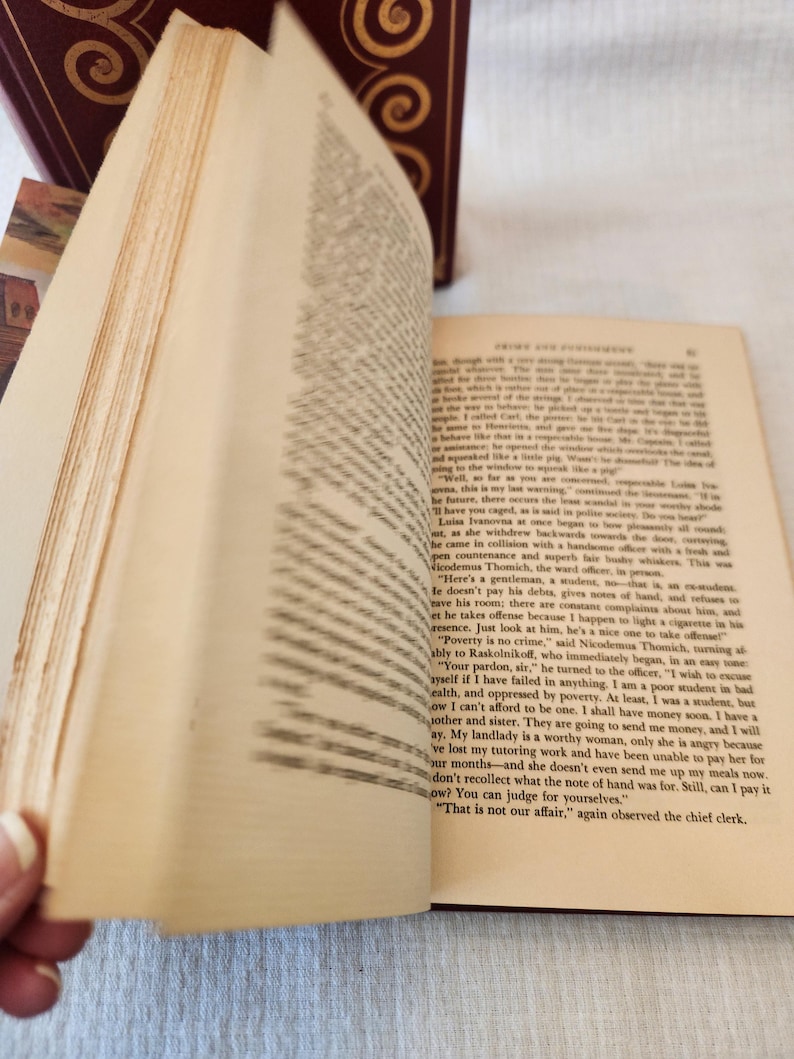 Peut inclure: Un livre reli&eacute; ouvert avec des pages couleur cr&egrave;me et du texte imprim&eacute; en noir. Le c&ocirc;t&eacute; gauche montre des pages floues en &eacute;ventail, tandis que le c&ocirc;t&eacute; droit affiche une page de texte claire tir&eacute;e de Crime et Ch&acirc;timent. Une couverture rouge fonc&eacute;.