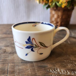 May include: White ceramic teacup with a blue rim and handle. The cup is decorated with a floral design in blue, yellow, and green. The cup is sitting on a wooden surface.