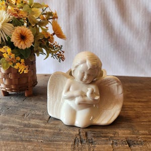 May include: A cream-colored ceramic angel figurine with outstretched wings, holding a small bird. The angel is seated, with detailed feather textures on the wings. A small basket of yellow and orange flowers is in the background.