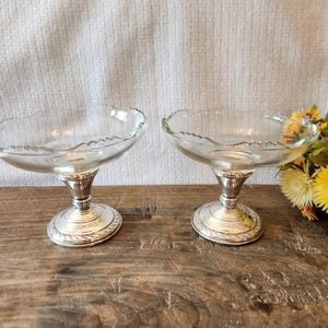 May include: Two clear glass dessert dishes with scalloped rims sit atop ornate silver-colored pedestals. The bases have a detailed, engraved design. The dishes are displayed on a wooden surface.