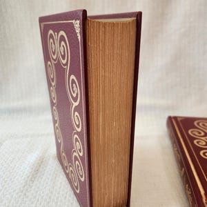 Peut inclure: Un livre &agrave; couverture rigide bordeaux avec une texture et une bordure dor&eacute;e en volutes. Les pages ont un aspect vieilli et beige. Un deuxi&egrave;me livre assorti est partiellement visible dans le coin inf&eacute;rieur droit.