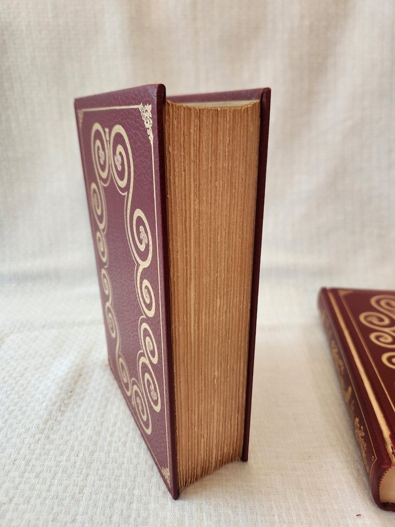 Peut inclure: Un livre &agrave; couverture rigide bordeaux avec une texture et une bordure dor&eacute;e en volutes. Les pages ont un aspect vieilli et beige. Un deuxi&egrave;me livre assorti est partiellement visible dans le coin inf&eacute;rieur droit.