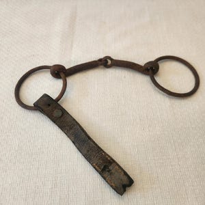 May include: An antique horse bit made of dark, aged metal with two large circular rings and a curved center piece. A worn, dark brown leather strap is attached to one of the rings, secured with a metal rivet. The bit shows signs of age and use.
