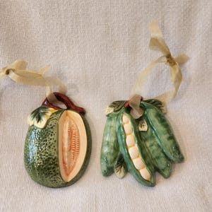 May include: Two ceramic vegetable ornaments. One is a green melon with a brown stem and beige interior. The other is a green pea pod with beige peas inside. Both have beige ribbon for hanging.