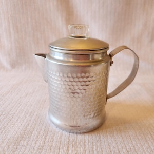 May include: A vintage silver-colored coffee pot with a textured, hammered metal body. The pot has a lid with a clear glass knob and a curved handle. The spout is on the left side. The pot is sitting on a cream-colored fabric.