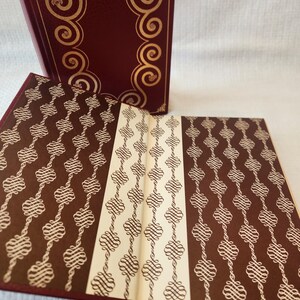 Peut inclure: Un livre reli&eacute; avec une couverture bordeaux profond orn&eacute;e de volutes dor&eacute;es en relief. Le livre ouvert pr&eacute;sente des pages de garde avec un motif r&eacute;p&eacute;titif de volutes en filigrane blanc sur fond brun fonc&eacute;, s&eacute;par&eacute;es par une bande centrale cr&egrave;me.