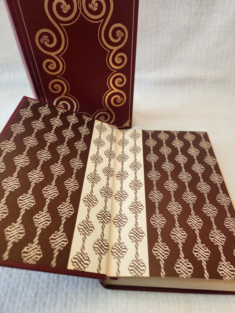 Peut inclure: Un livre reli&eacute; avec une couverture bordeaux profond orn&eacute;e de volutes dor&eacute;es en relief. Le livre ouvert pr&eacute;sente des pages de garde avec un motif r&eacute;p&eacute;titif de volutes en filigrane blanc sur fond brun fonc&eacute;, s&eacute;par&eacute;es par une bande centrale cr&egrave;me.