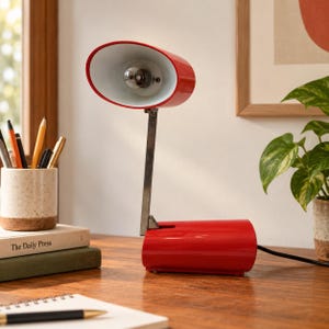 May include: A bright red desk lamp with an oval-shaped head and a white interior. The lamp features a metallic silver arm connected to a cylindrical red base. A black power cord extends from the back of the base, providing a functional lighting solution.