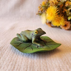 May include: A ceramic frog figurine with yellow eyes and green spots, resting on a green leaf. The frog is detailed with textured skin and sits atop a leaf with visible veins. A bouquet of yellow flowers is in the background.