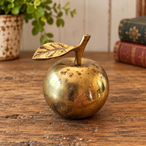 May include: A polished brass apple figurine featuring a smooth, rounded body and a single attached leaf on the stem. The metal surface displays a warm, golden tone with subtle dark patina spots and visible texture on the leaf veins.