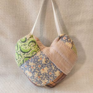 May include: A handmade heart-shaped ornament composed of various fabric patches. The patches feature patterns in green, blue, and pink, with a white ribbon for hanging. The ornament is a patchwork design.