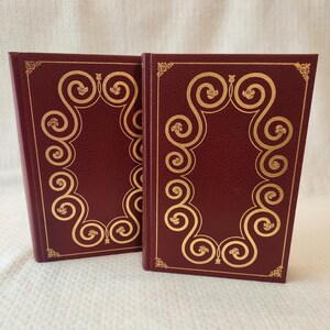 Peut inclure: Deux livres &agrave; couverture rigide assortis avec des couvertures bordeaux profond pr&eacute;sentant une surface textur&eacute;e. Chaque couverture est orn&eacute;e d'un cadre dor&eacute; en feuille d'or compos&eacute; de volutes complexes et de petits motifs floraux aux coins.