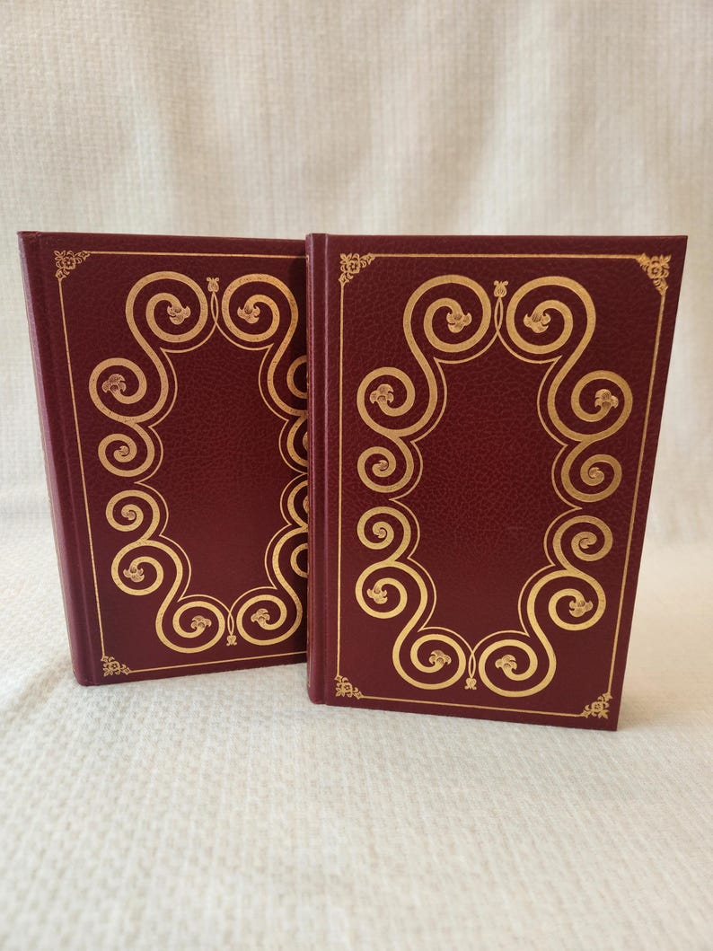 Peut inclure: Deux livres &agrave; couverture rigide assortis avec des couvertures bordeaux profond pr&eacute;sentant une surface textur&eacute;e. Chaque couverture est orn&eacute;e d'un cadre dor&eacute; en feuille d'or compos&eacute; de volutes complexes et de petits motifs floraux aux coins.