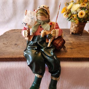May include: A whimsical frog figurine dressed as a fisherman, wearing a red and white checkered shirt, green waders, and a fishing hat. The frog holds a fishing rod, net, and tackle box. A small basket of yellow flowers sits in the background.