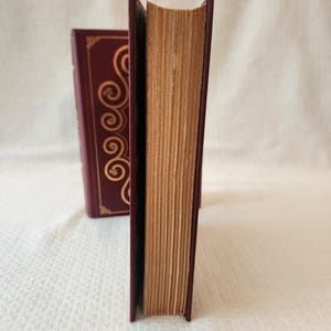 Peut inclure: Deux livres &agrave; couverture rigide bordeaux profond avec des motifs de volutes dor&eacute;s sur les tranches. Les livres sont plac&eacute;s verticalement, r&eacute;v&eacute;lant les bords textur&eacute;s, vieillis et brun clair des pages en papier entre les couvertures sombres.